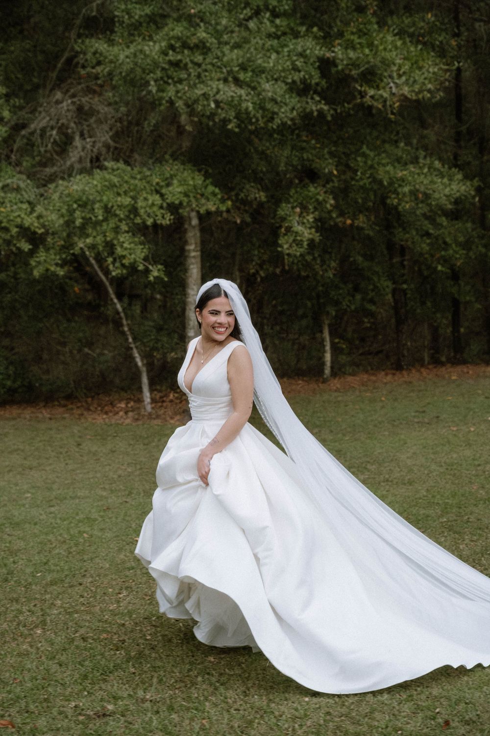 A bride in a flowing white wedding dress and veil poses playfully on a grassy lawn surrounded by trees.