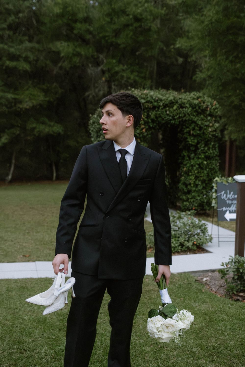 Groom in black suit walking through garden setting.