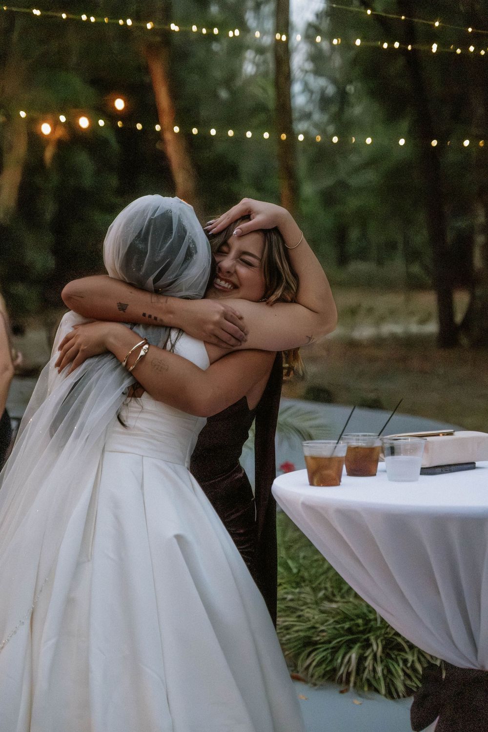 Two people share an emotional embrace at an outdoor wedding reception with string lights and white tablecloths.