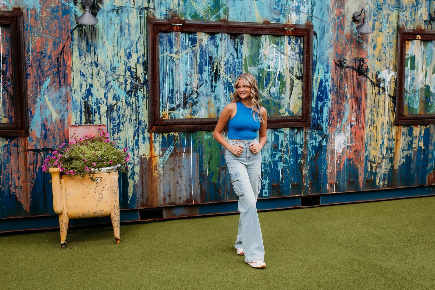 A figure poses with an empty picture frame against a rustic blue wall with peeling paint and artificial turf flooring.