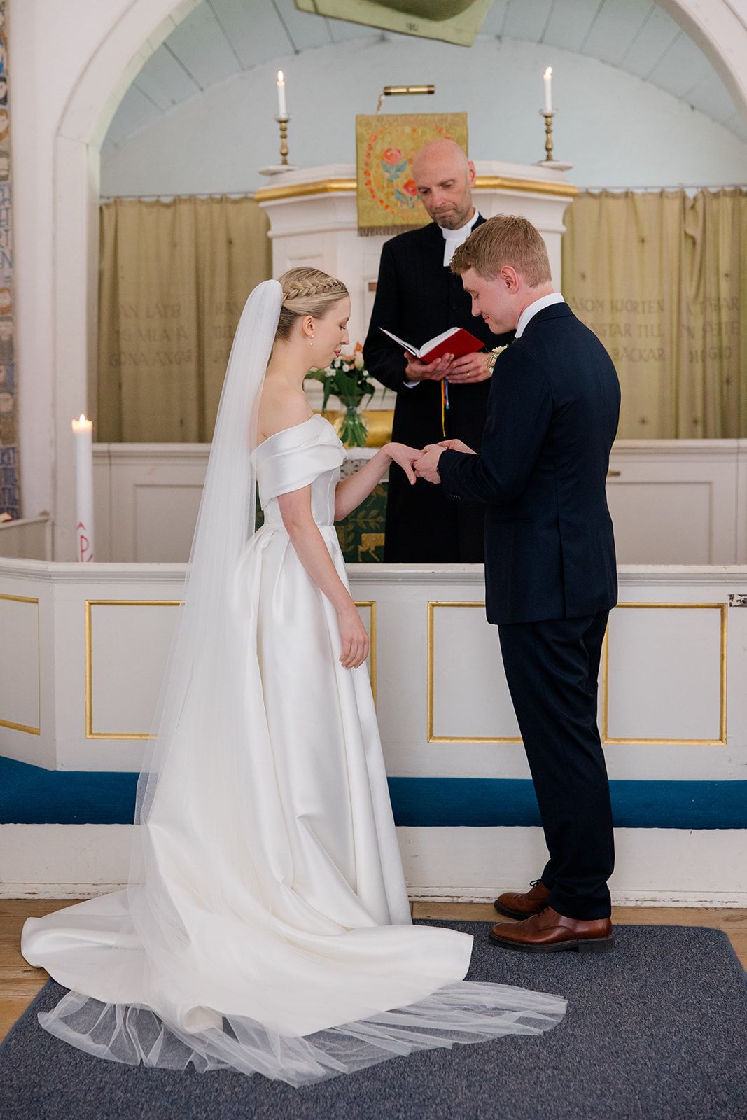 A wedding ceremony takes place in a church with a bride in a flowing white dress exchanging rings at the altar.