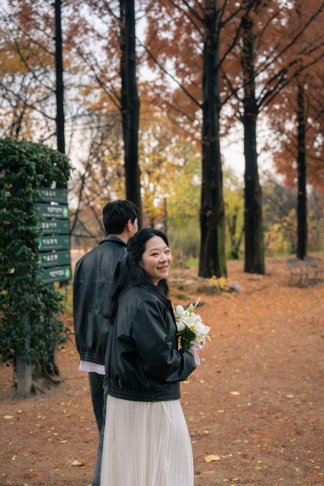 A person in a black jacket and white skirt stands among autumn trees with fallen leaves in a park setting.