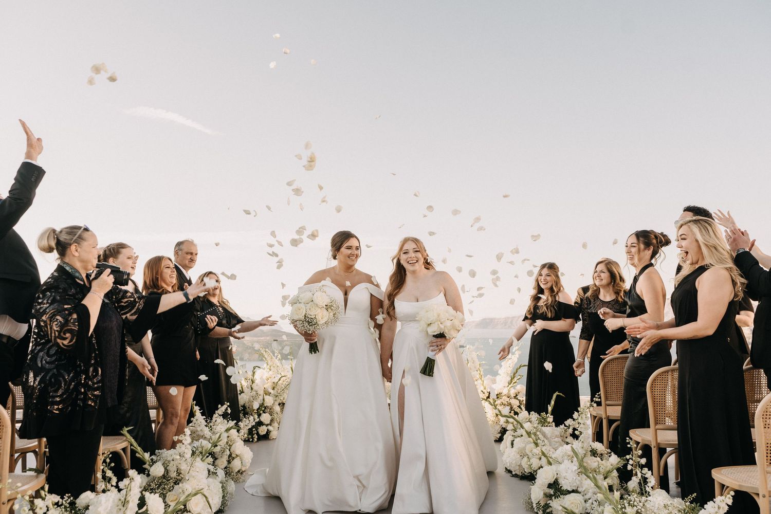 Wedding guests gather around an elegant outdoor ceremony setup with white floral arrangements and decorations.