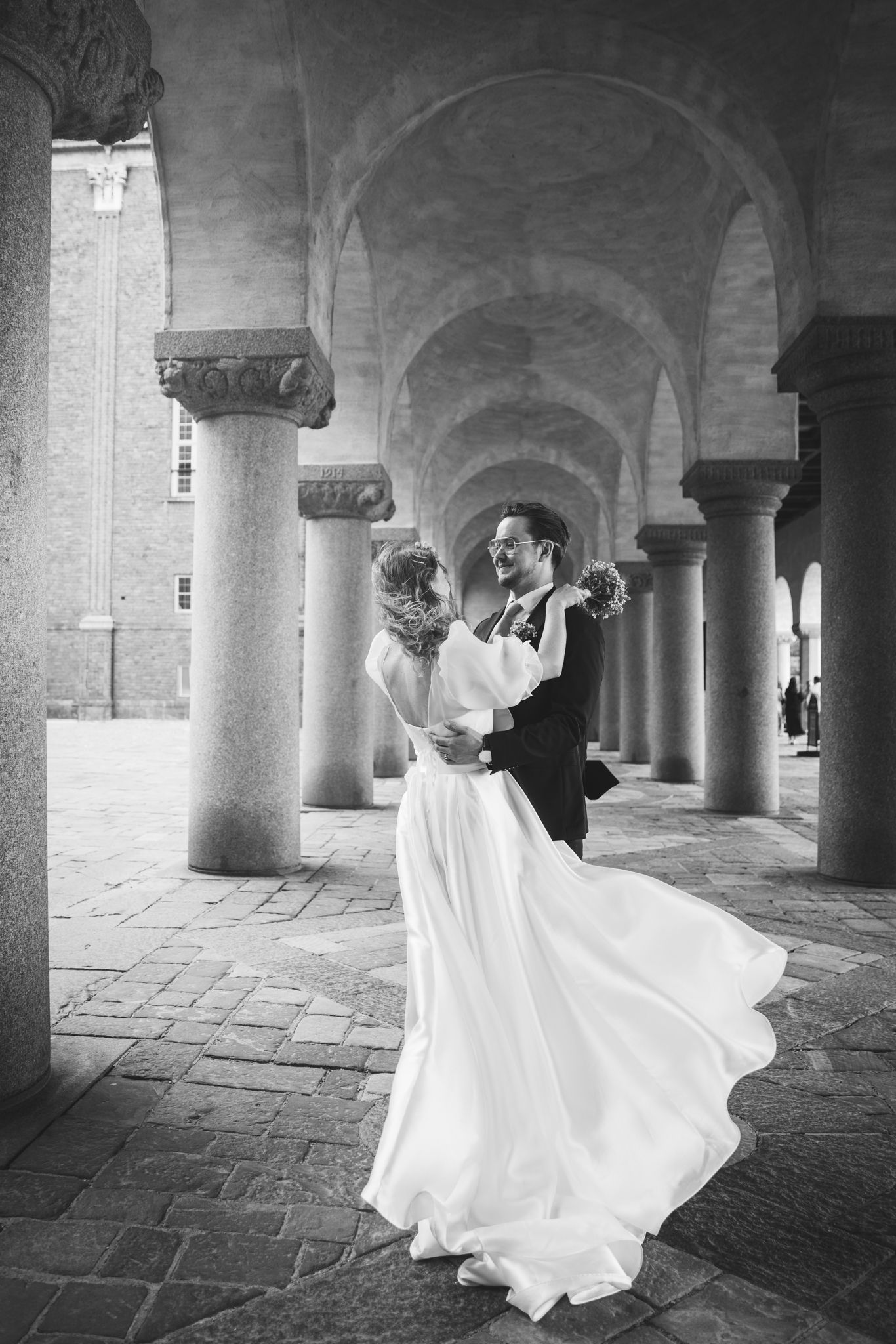 A romantic black and white photo of a couple dancing in an elegant colonnade with flowing white dress and classic architecture.