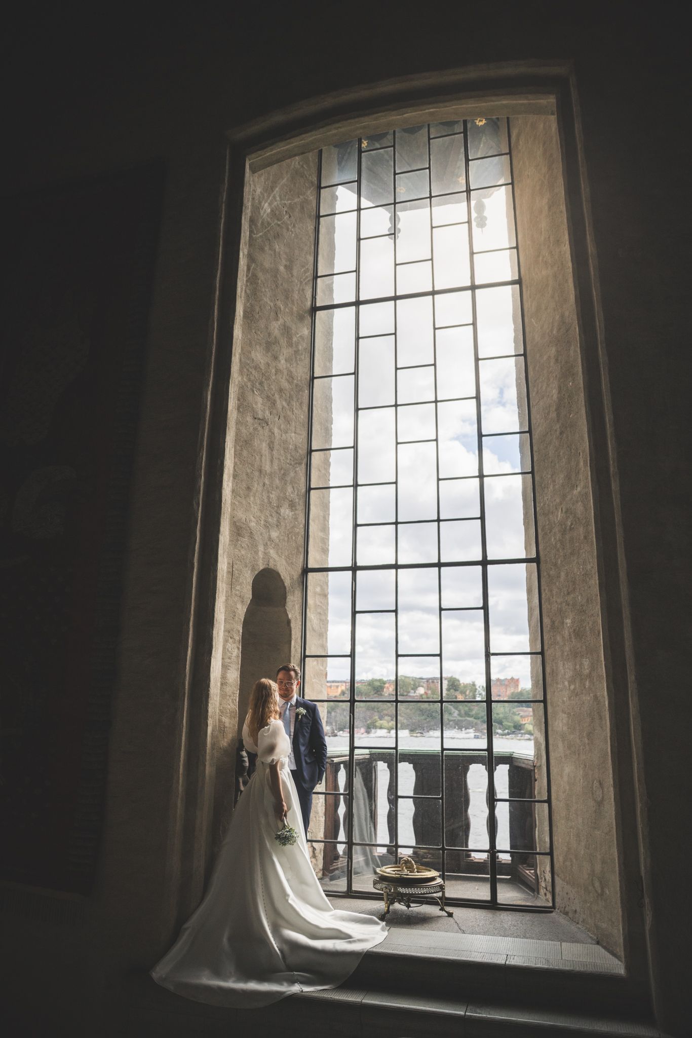 Couple stands in elegant arched window alcove with dramatic lighting and geometric glass panes in historic building.