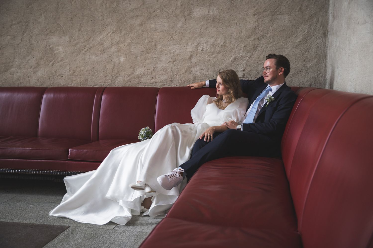 A couple in wedding attire relaxes together on a burgundy leather couch in an industrial setting.