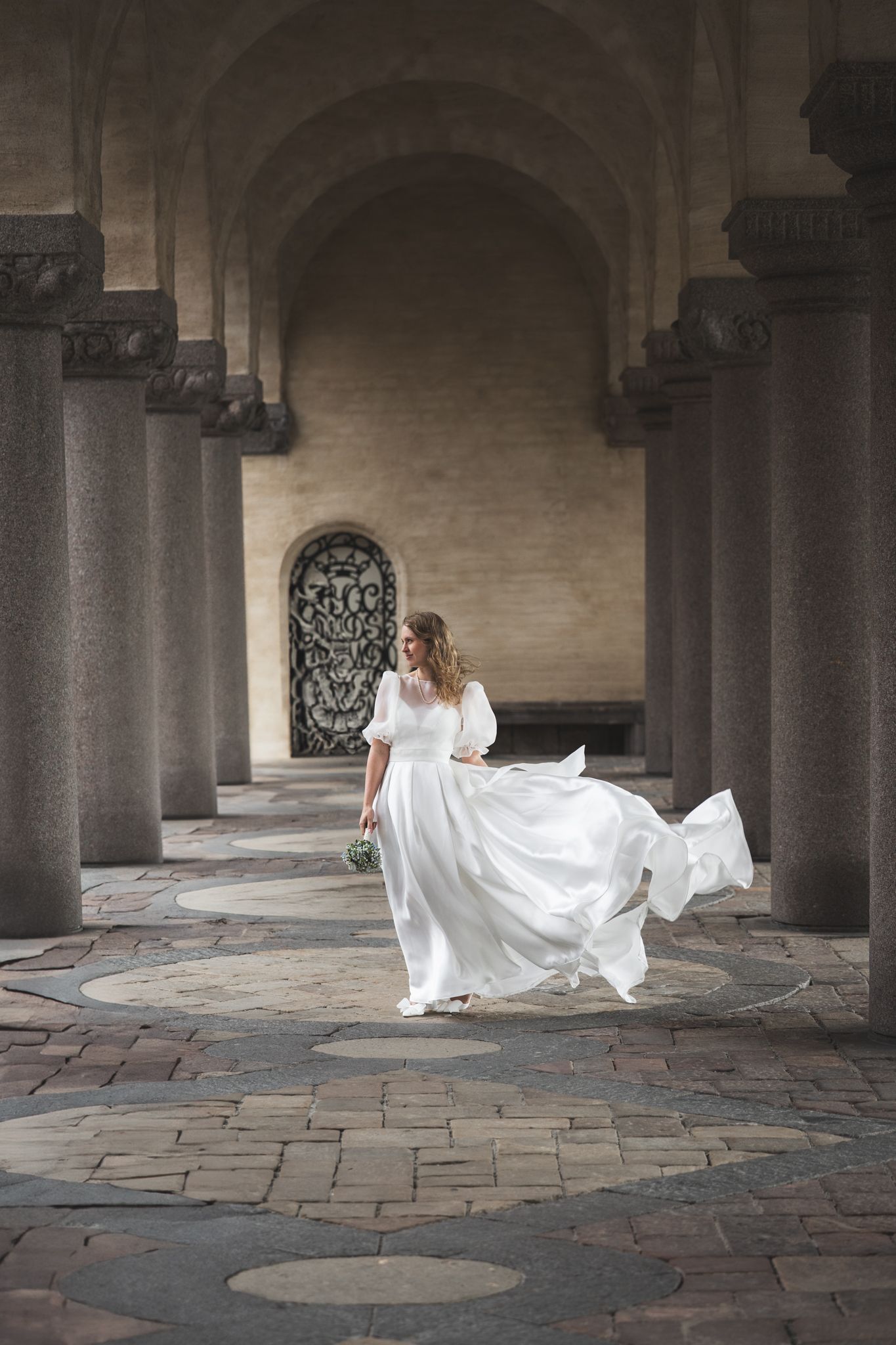A flowing white wedding dress twirls through a series of stone archways in a historic courtyard setting.