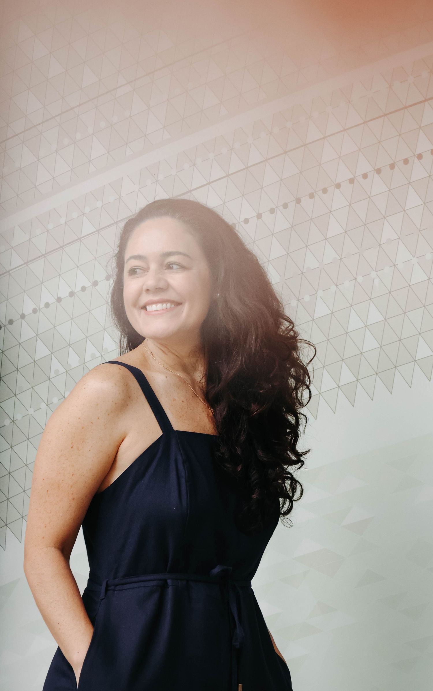 A happy woman with long curly hair wearing a navy dress smiles outdoors against a geometric patterned wall.