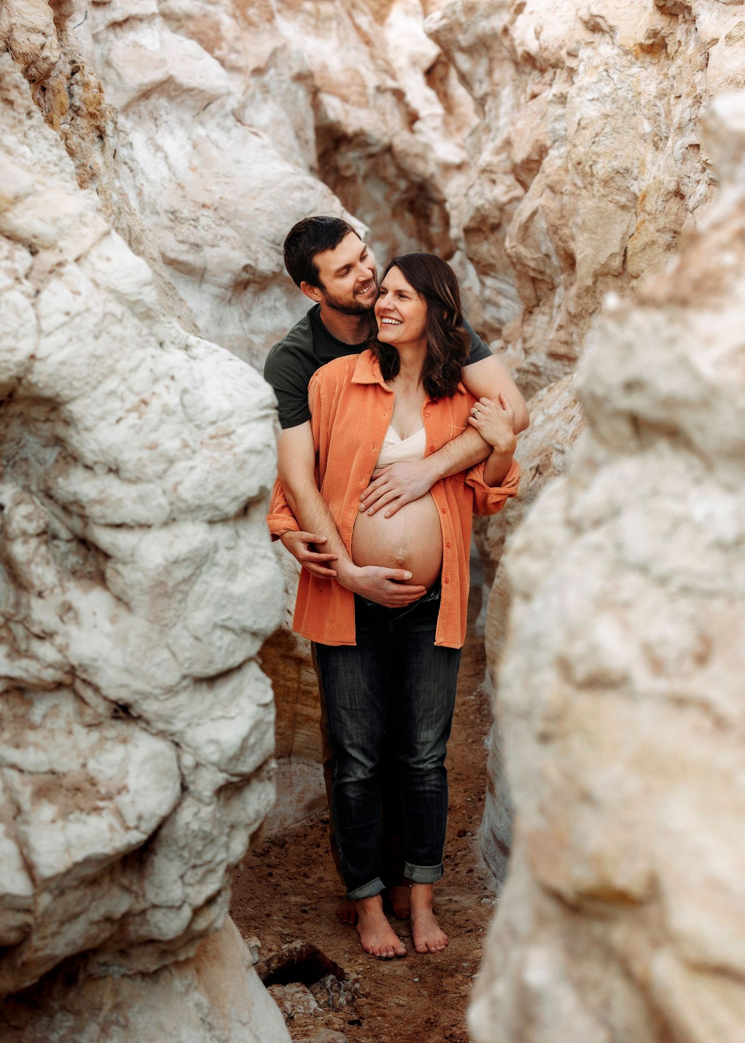 Couple embracing in rocky canyon, man holding pregnant woman's belly, smiling together.