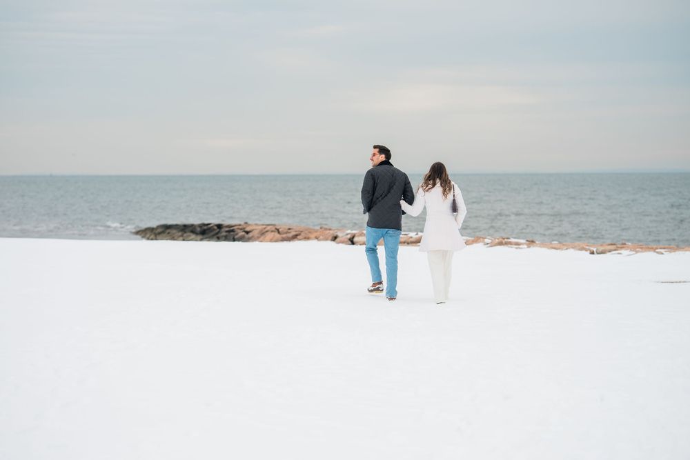 Two people walk together on a snowy beach with ocean waves and cloudy skies in the background.