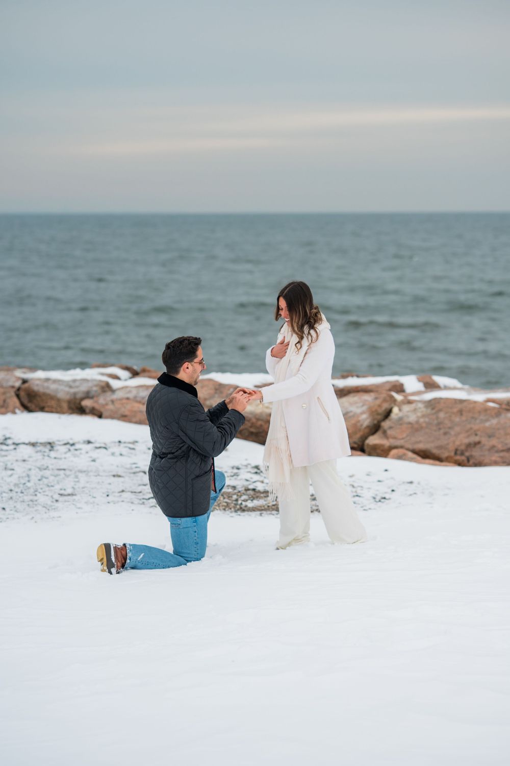 A couple sits together on a snowy beach with large rocks and ocean waves in the background.