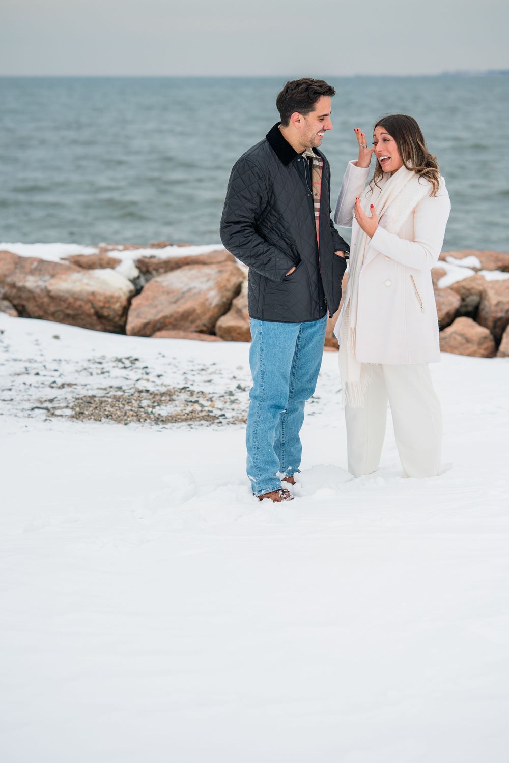 A couple shares a romantic moment on a snowy beach with rocky shoreline and ocean waves in the background.