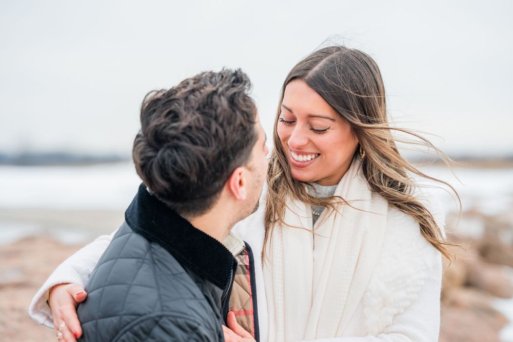 Couple in winter coats embracing and laughing together on a snowy beach.