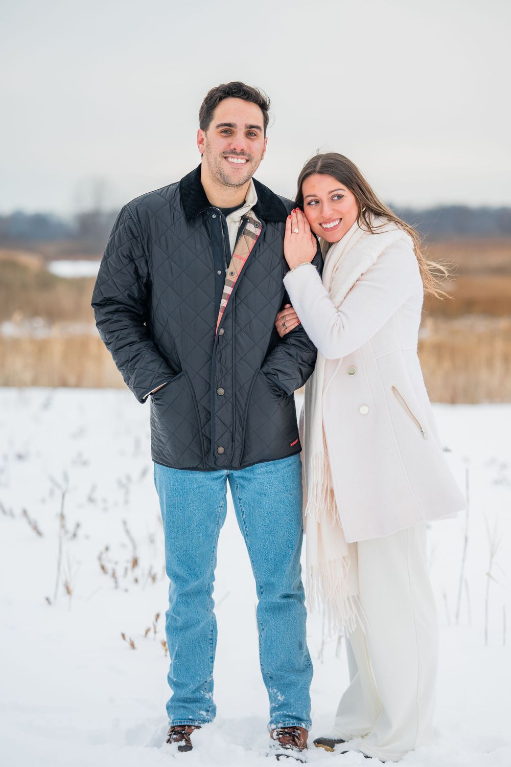 A couple poses together in a snowy winter field wearing casual outerwear.