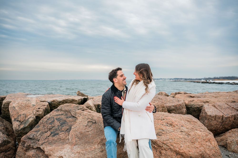 A couple shares a romantic moment on coastal rocks overlooking the ocean on a bright sunny day.