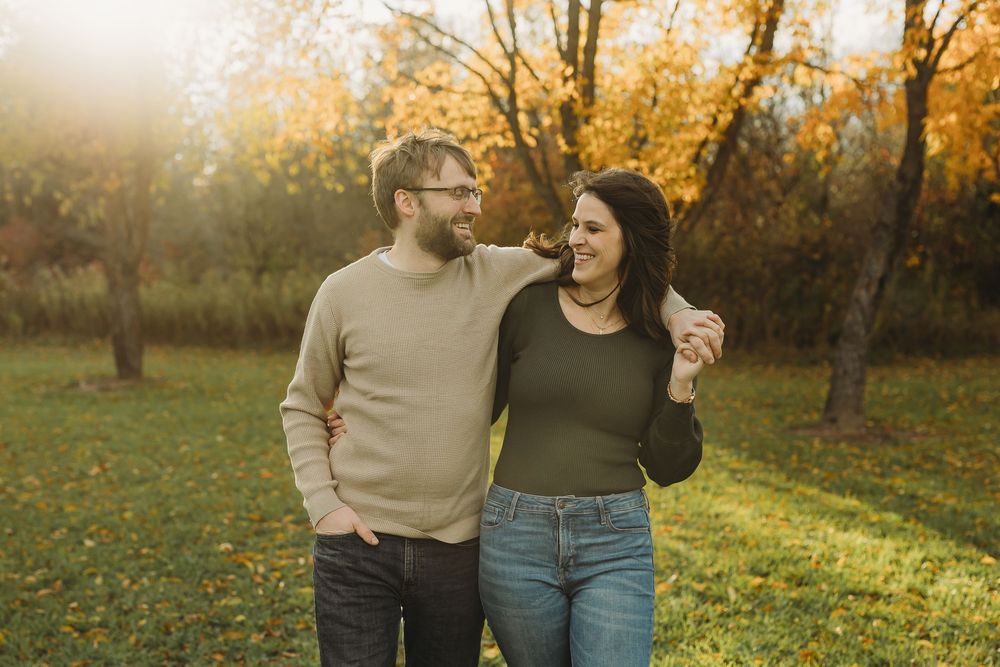 Couple walking together through an autumn orchard at golden hour with fallen leaves on the ground.