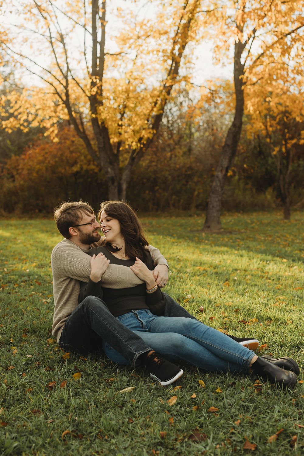 A couple lies together on autumn grass surrounded by fallen leaves in a romantic outdoor setting.