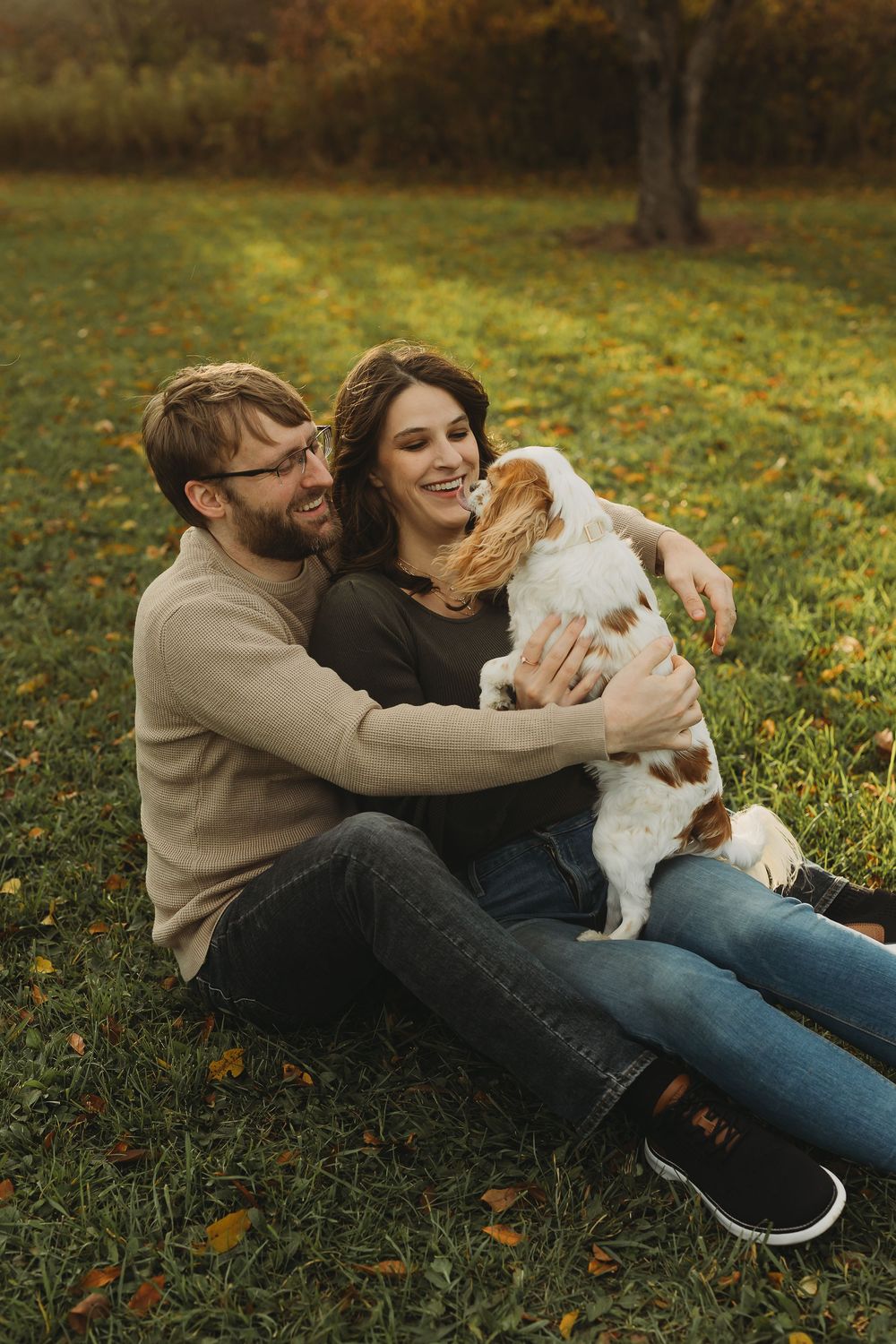 A couple sits together on autumn grass with their small white dog during a cozy outdoor photoshoot.