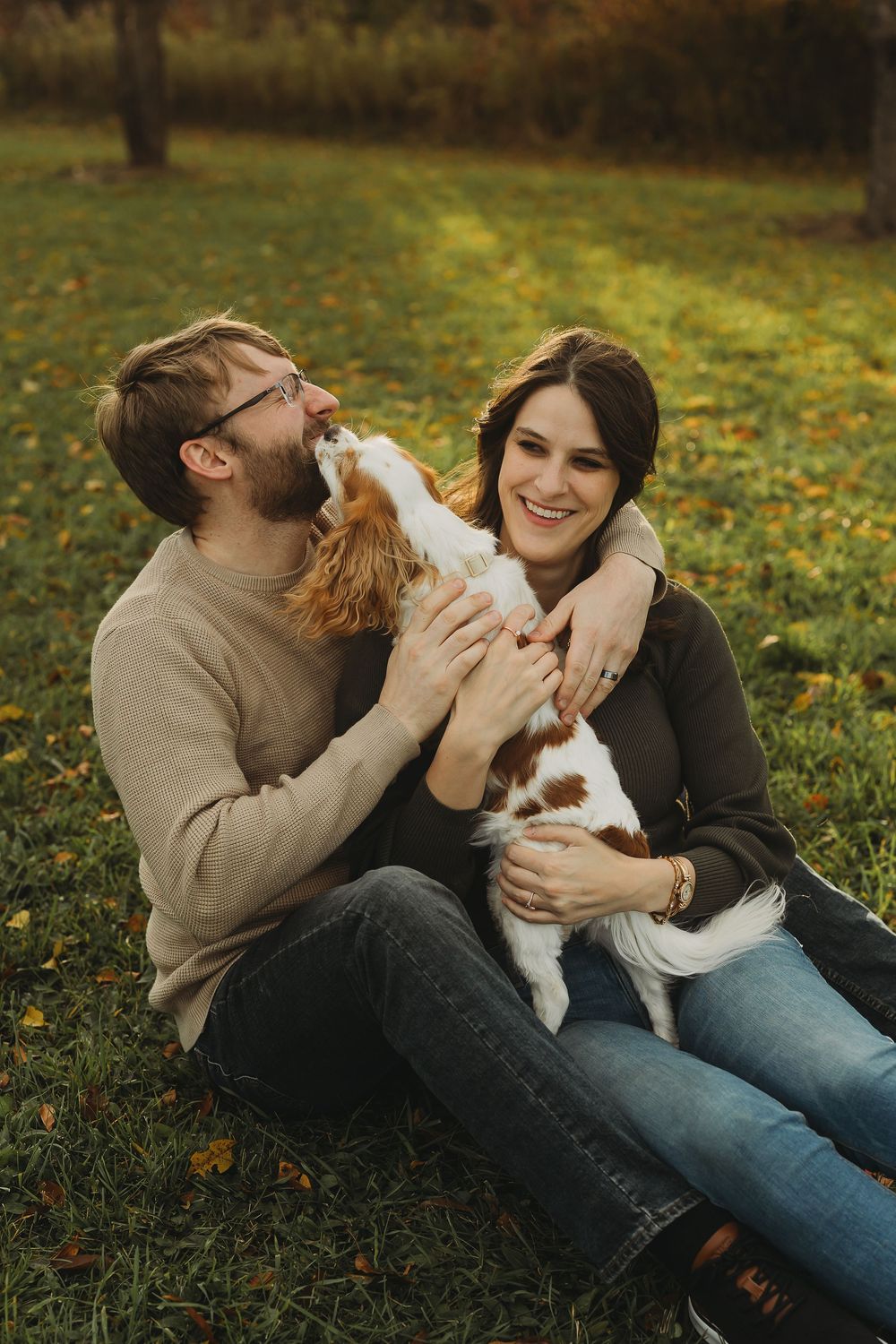 A couple sits on grass with their small white and brown dog in a series of playful autumn photos.