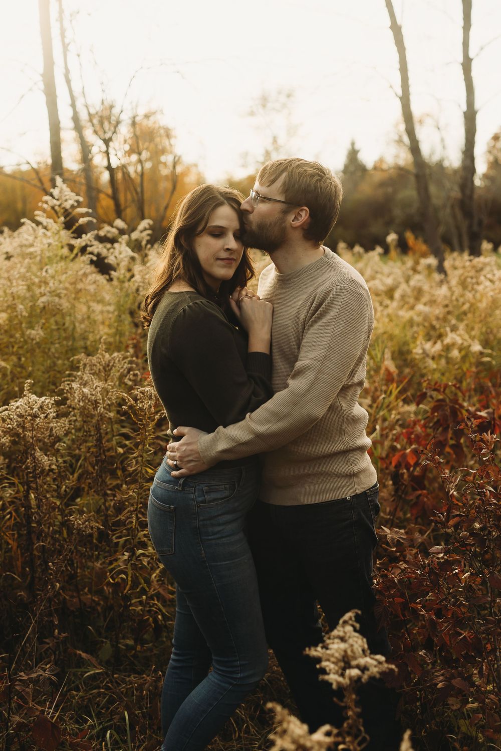 A romantic couple embraces in a golden field with autumn foliage at sunset.
