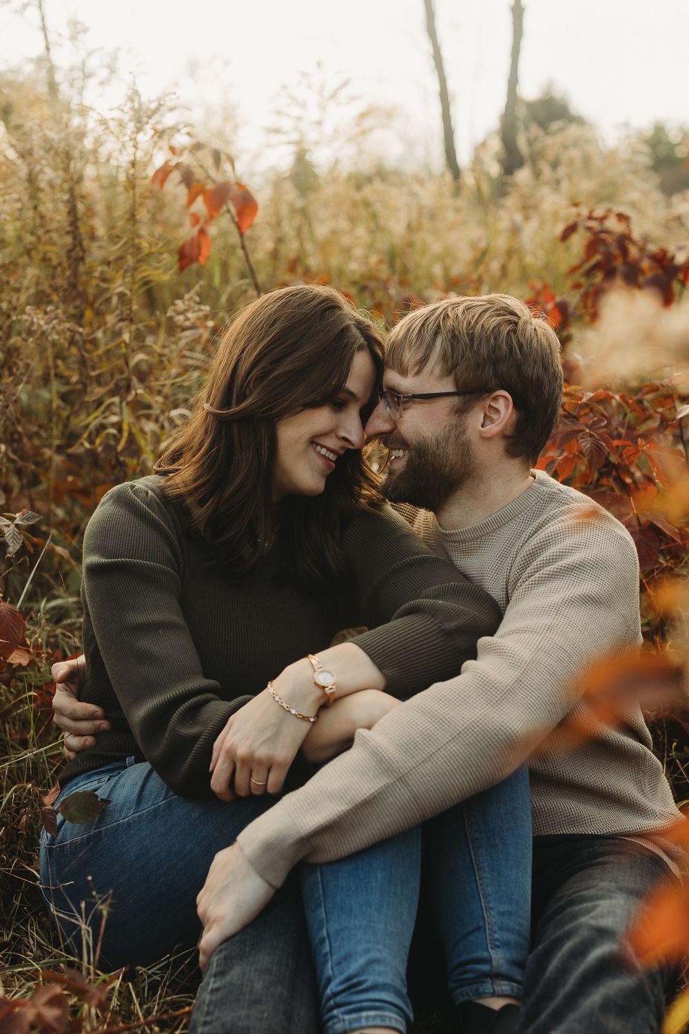 A couple shares tender moments in autumn foliage wearing casual sweaters and jeans while sitting outdoors.