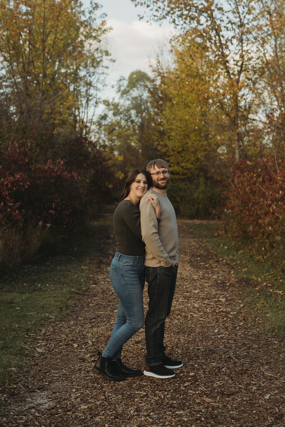 A couple shares an intimate moment while walking along a leaf-covered trail surrounded by autumn trees.
