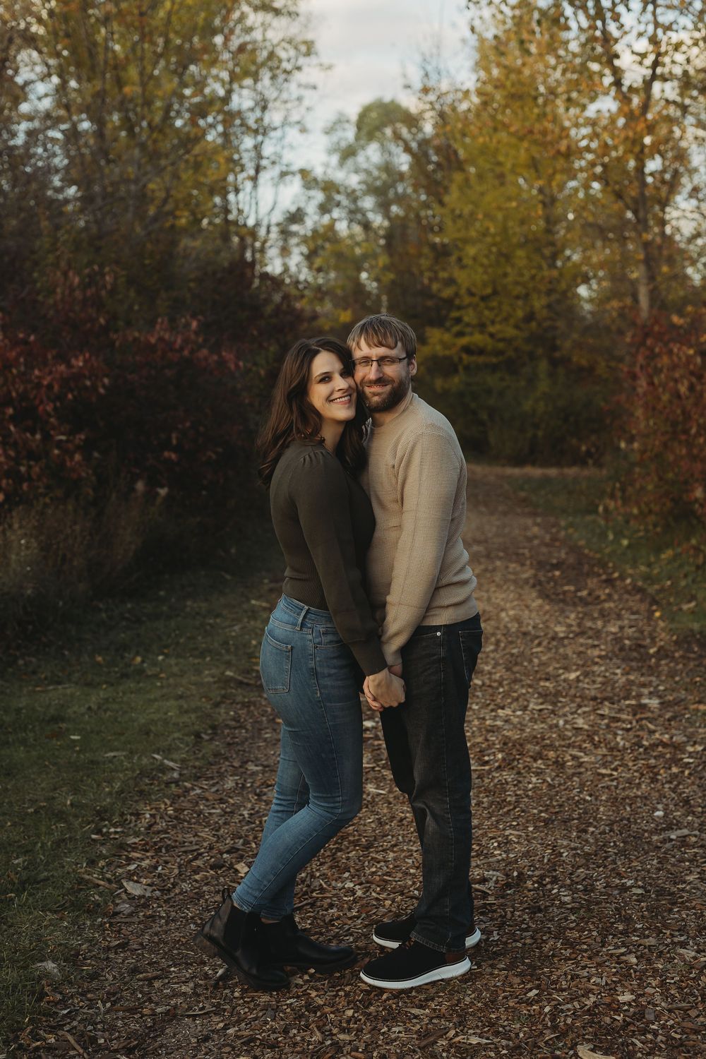 A couple in casual attire embraces on a leaf-covered path during autumn with trees in the background.