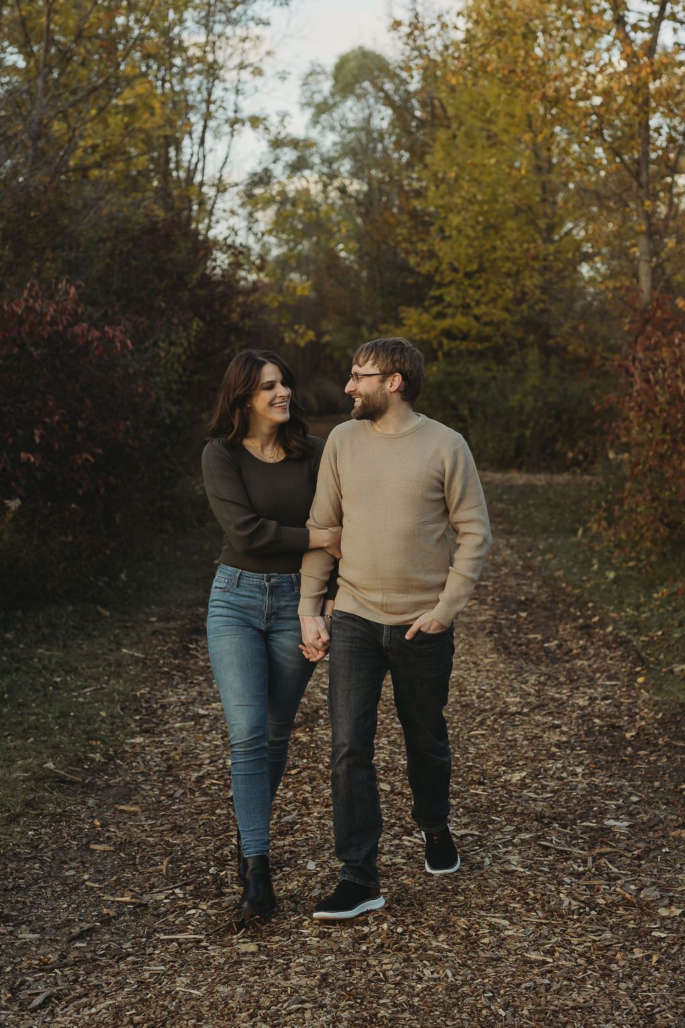 A couple walks together on a leaf-covered path during autumn with trees in the background.