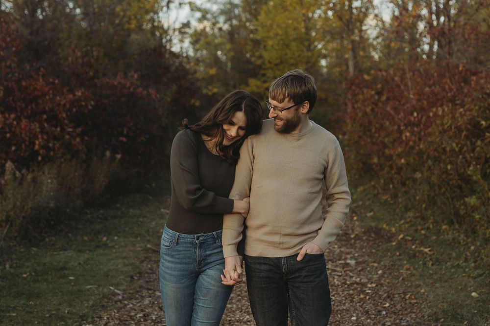 A couple walks along an autumn path surrounded by trees with red and orange foliage in the background.