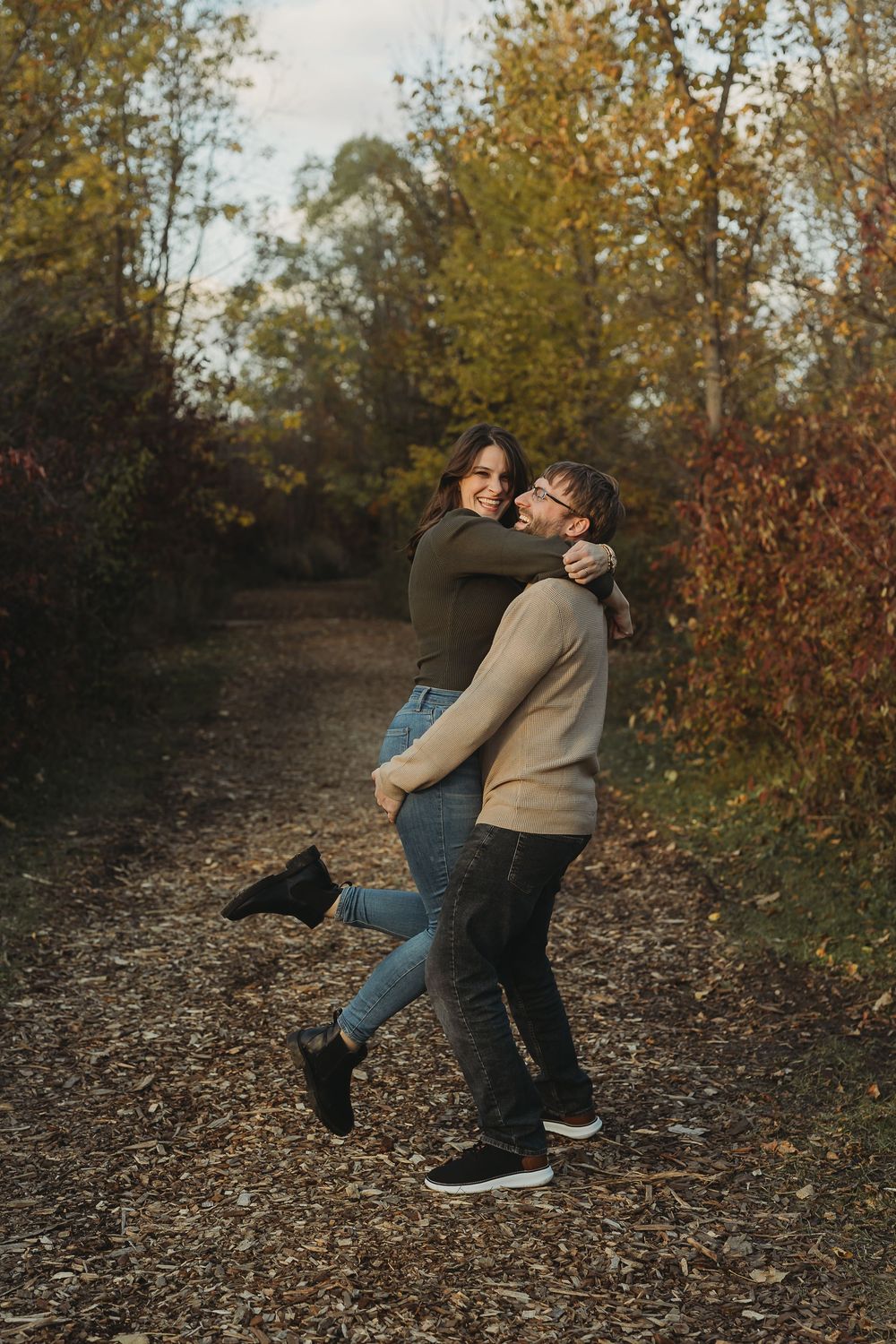 A couple in casual fall attire playfully spins and dances together on a leaf-covered forest path during autumn.