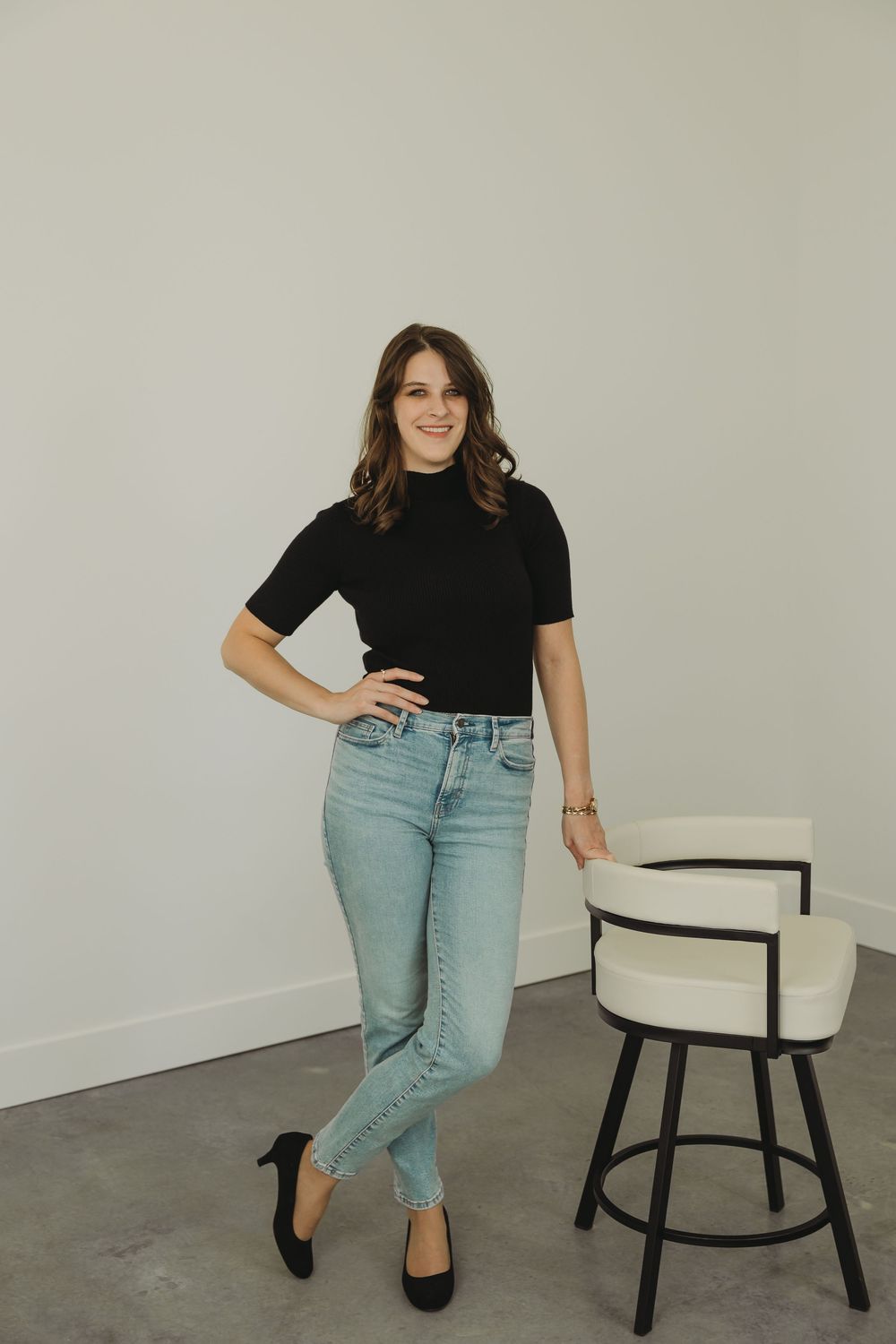 Woman in black t-shirt and light jeans poses casually with modern white and black accent chair in studio setting.