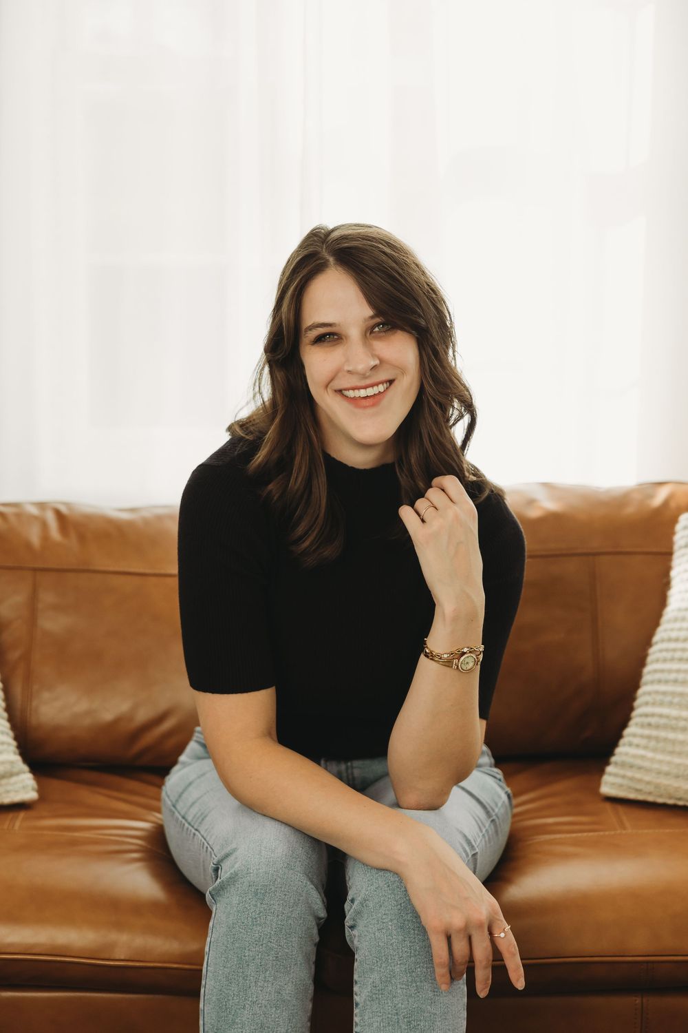A person wearing a black sweater sits casually on a brown leather couch in a bright, minimalist room.