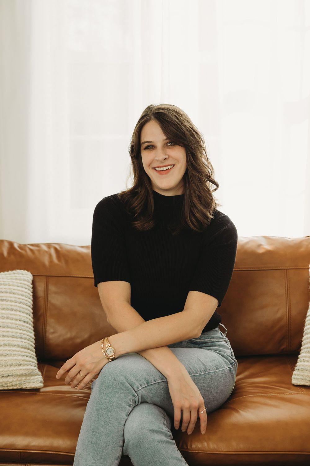 Casual portrait on a brown leather couch wearing a black turtleneck and light denim jeans in a bright room.