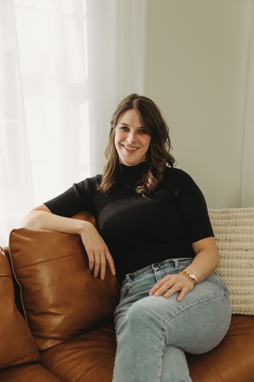 Casual portrait on brown leather couch wearing black top and light denim jeans in natural window light.