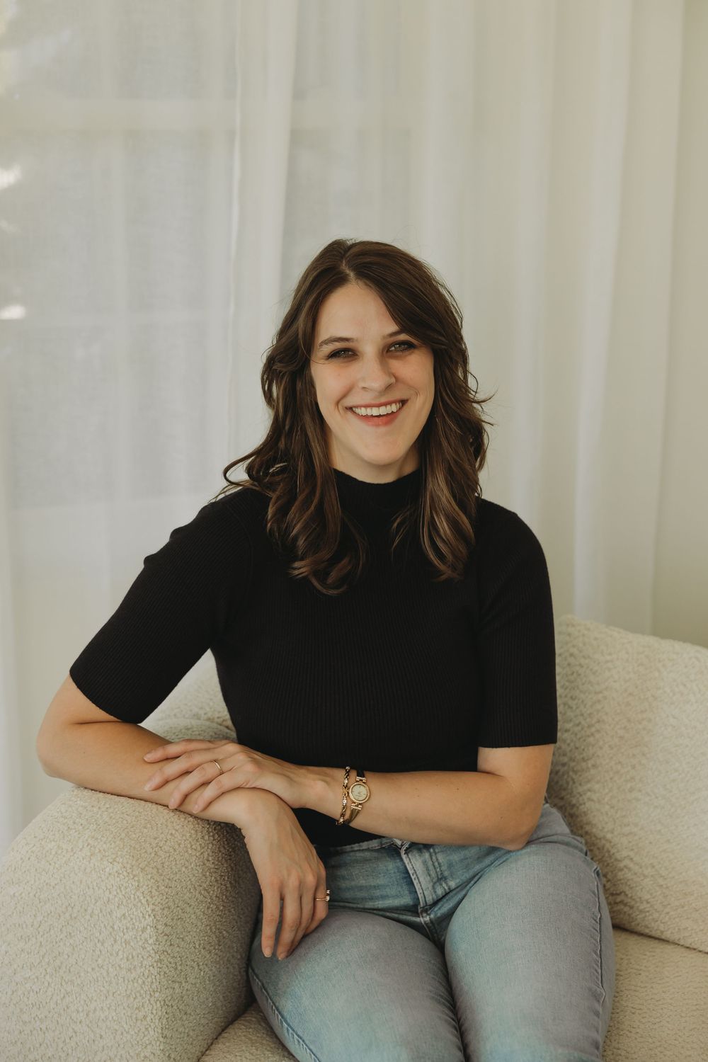 A person in a black short-sleeved top and jeans sits casually on a beige couch against a light curtained background.