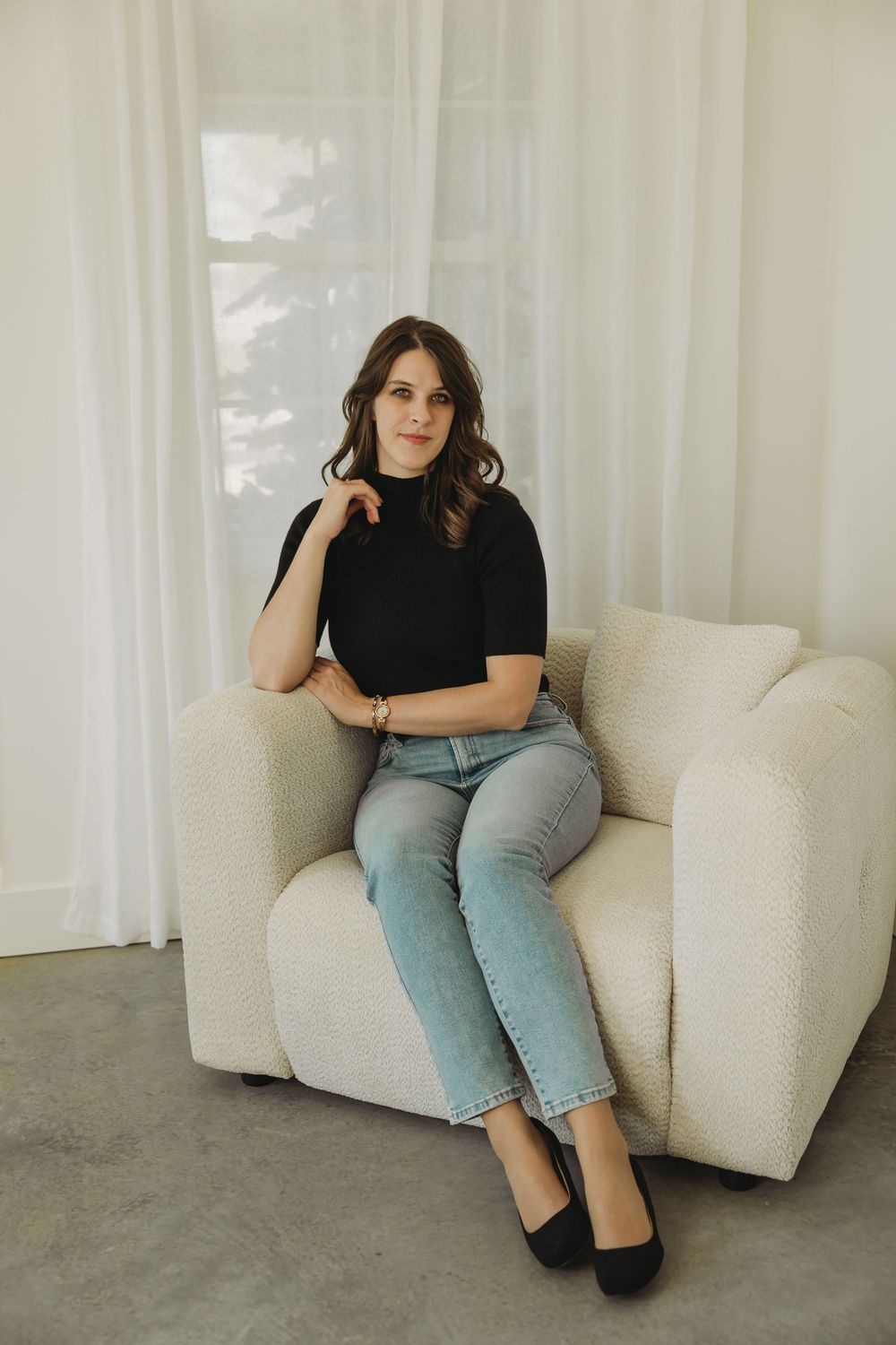 A person in a black top and light jeans sits casually in different positions on a cream-colored armchair.