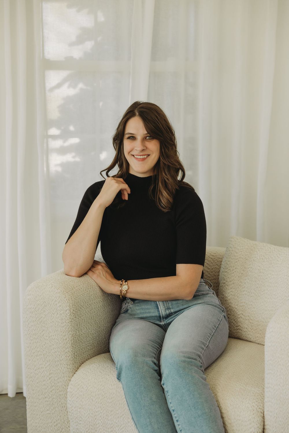 Person in black shirt and jeans sits casually on cream-colored couch in bright room with sheer curtains.