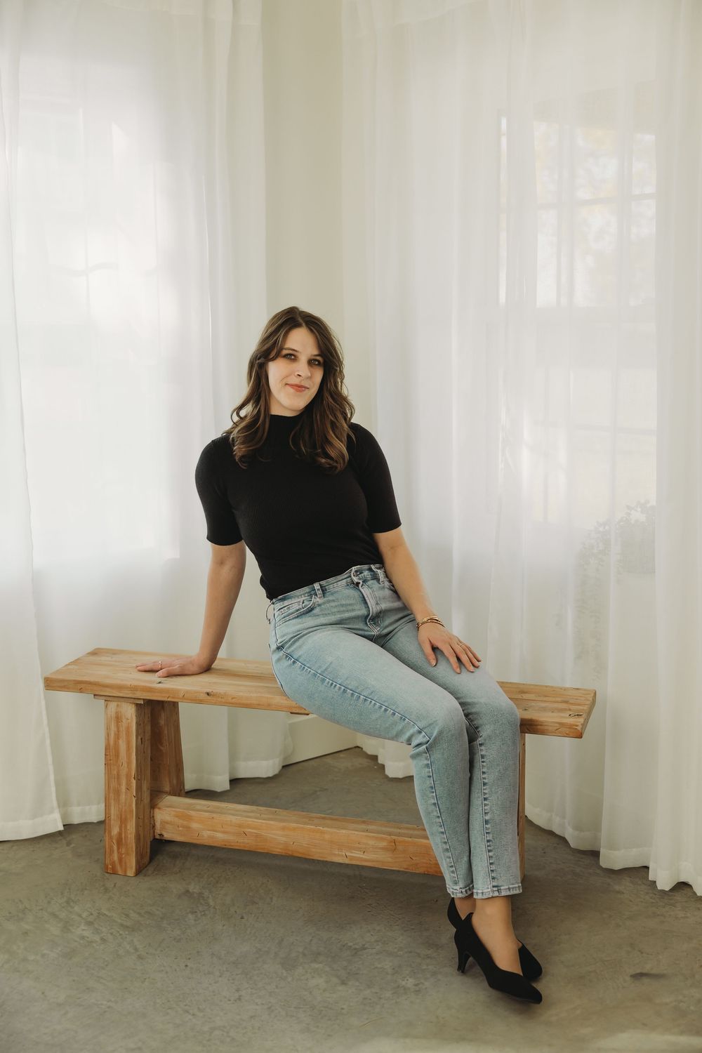 A person in a black top and light blue jeans poses on a wooden bench against a white curtained backdrop.