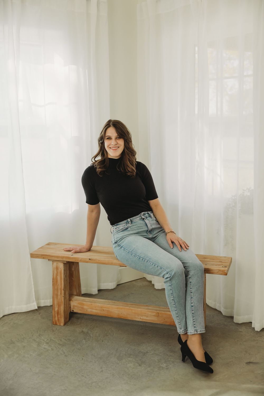 A person in a black top and light jeans sits casually on a wooden bench against a white curtained backdrop.