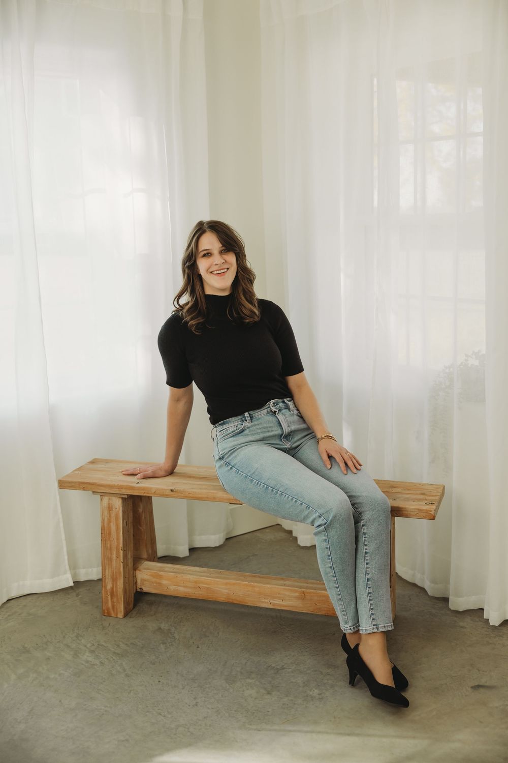A person in a black t-shirt and light jeans sits casually on a wooden bench against a white curtained background.