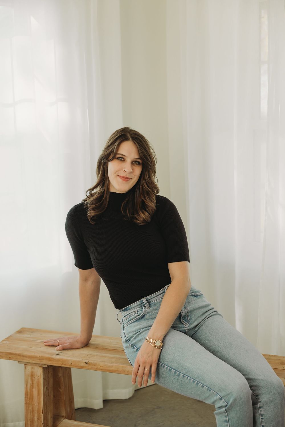 A person in a black t-shirt and light jeans sits casually on a wooden bench against a white background.