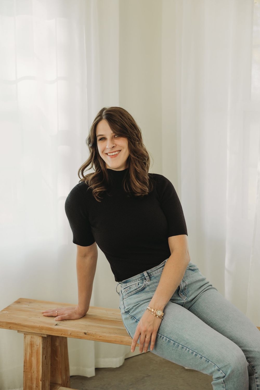 Person in black turtleneck and light jeans poses casually on wooden bench in bright white studio setting.