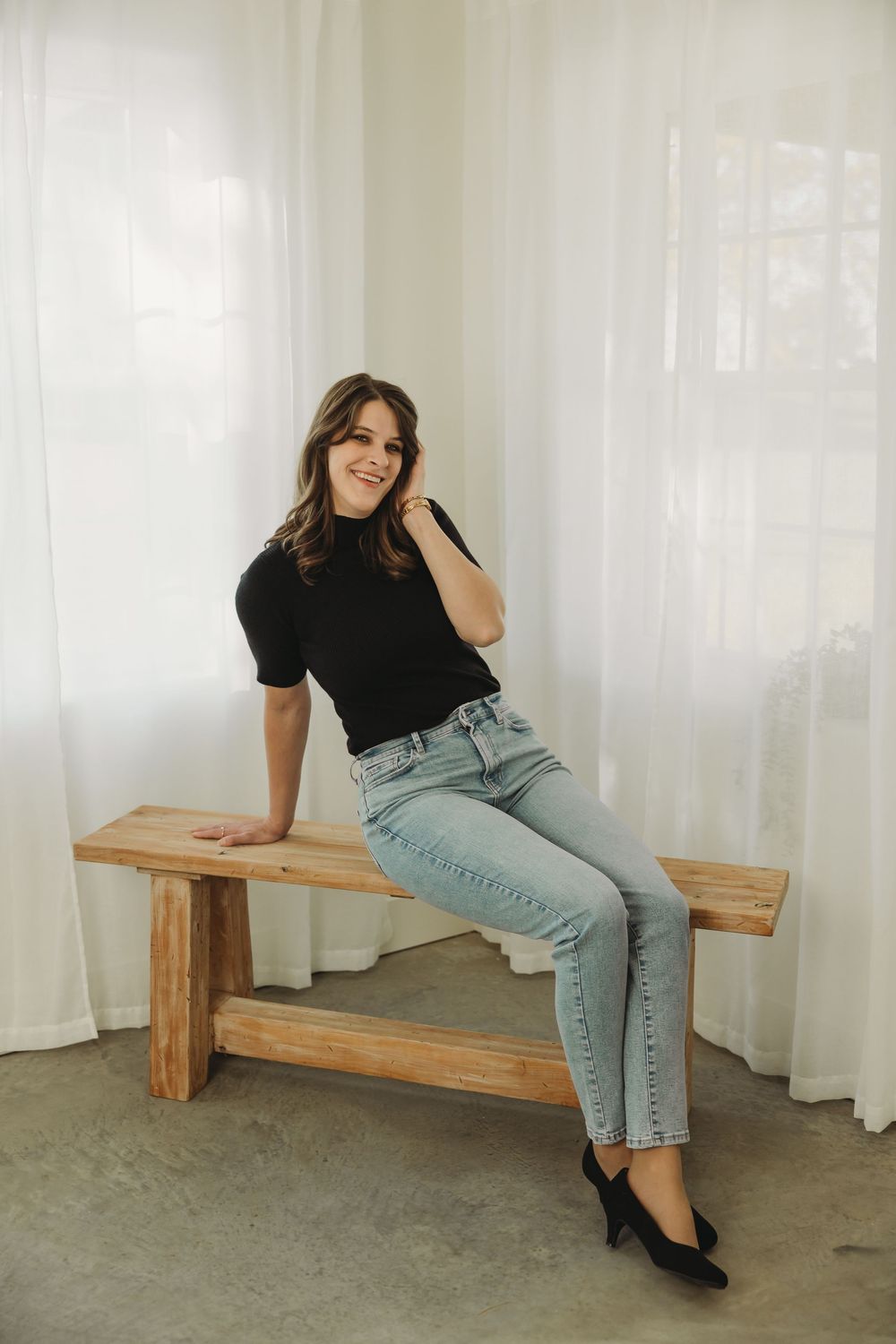 A person in a black top and light wash jeans poses casually on a wooden bench against a white curtained backdrop.