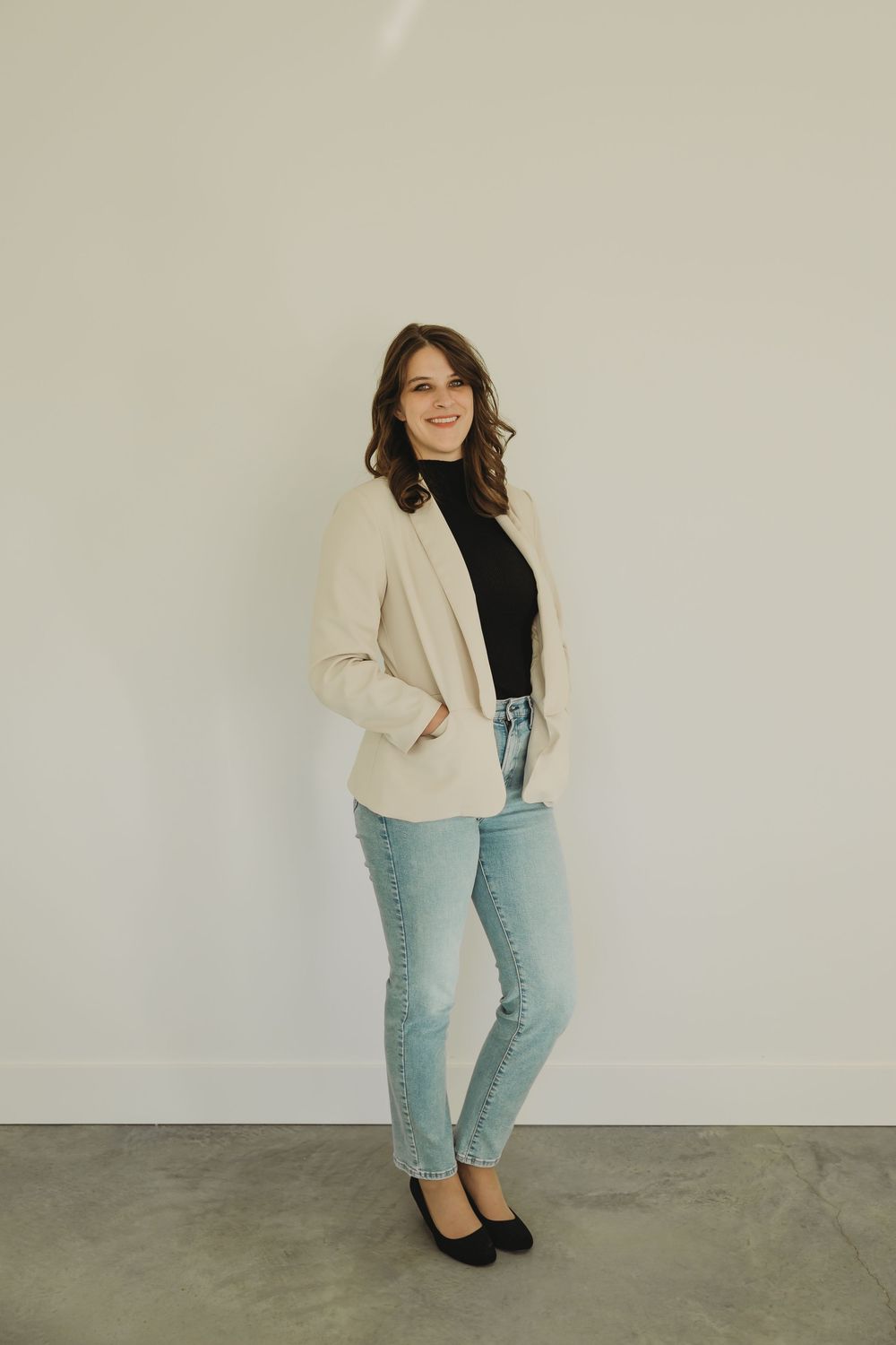 A person wearing a cream blazer, black top, light blue jeans and black heels poses against a white wall.