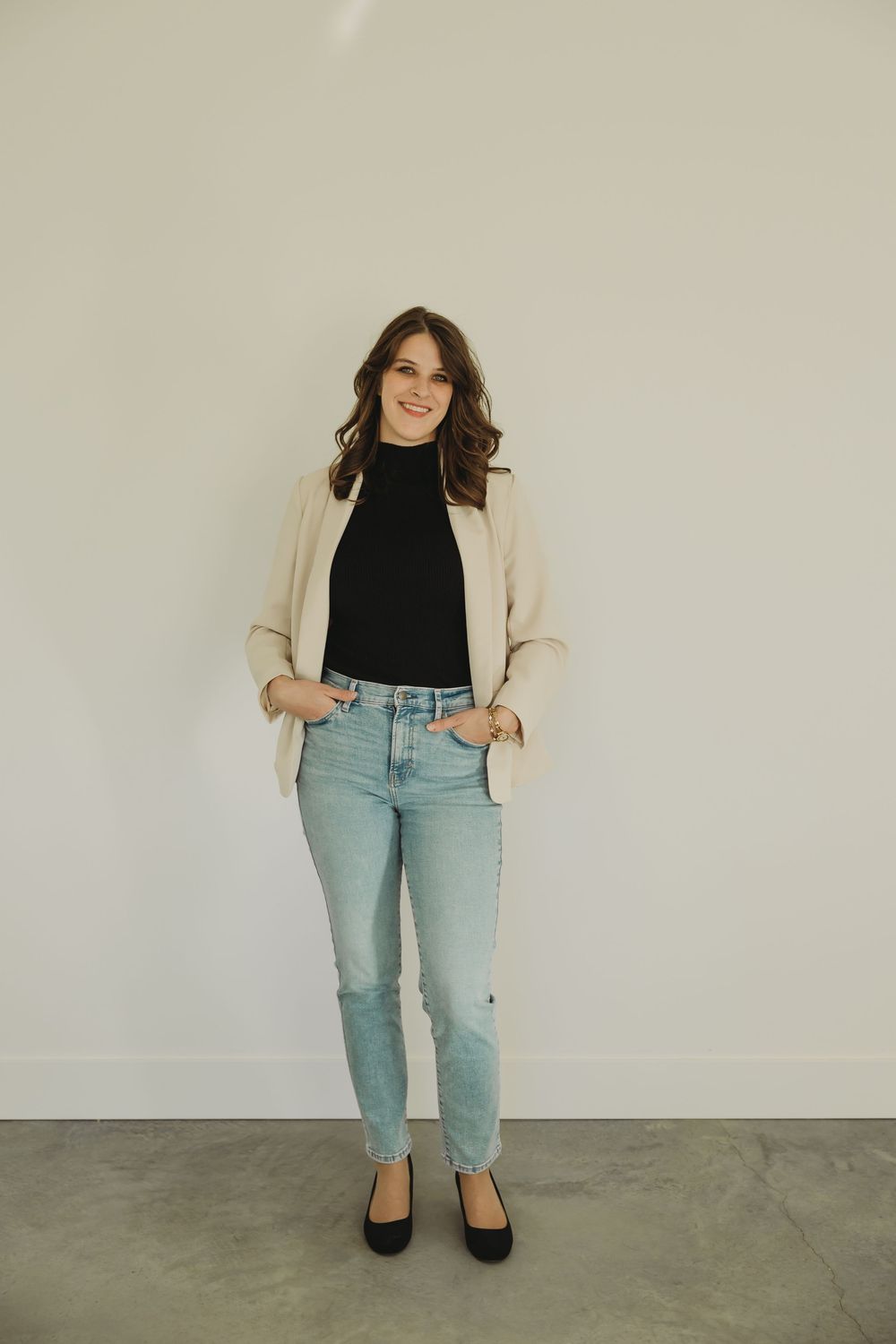 Person wearing a cream cardigan, black top, light wash jeans and black flats poses against a white wall.