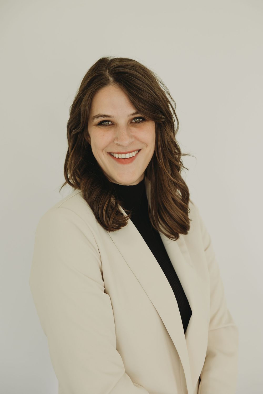 Professional studio portrait showing someone wearing a cream blazer and black top with a warm smile against neutral backdrop.