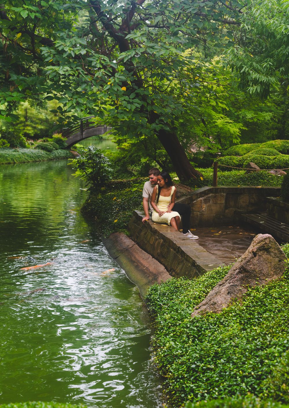 A peaceful lake surrounded by lush greenery with a stone retaining wall and people relaxing by the water's edge.