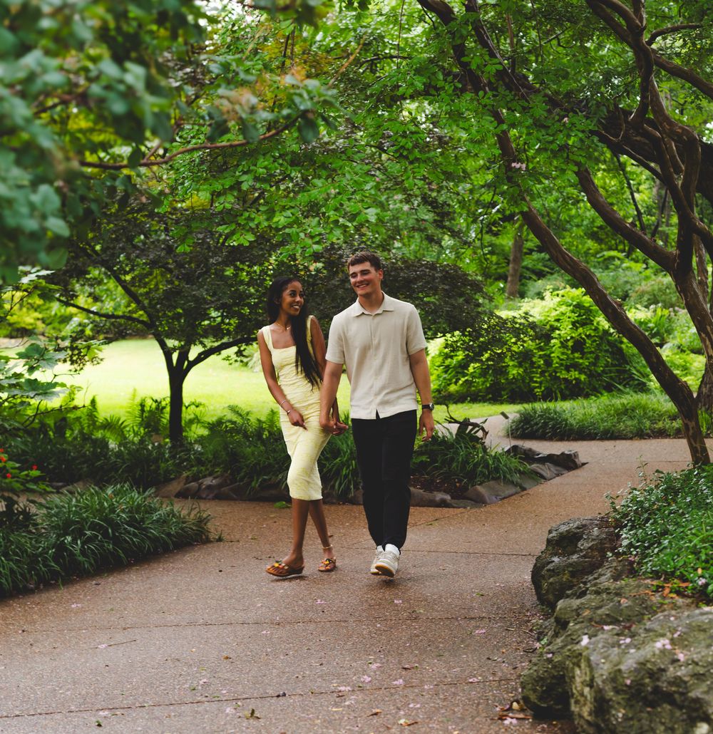Couple walking together on a winding garden path surrounded by lush green trees and landscaping.