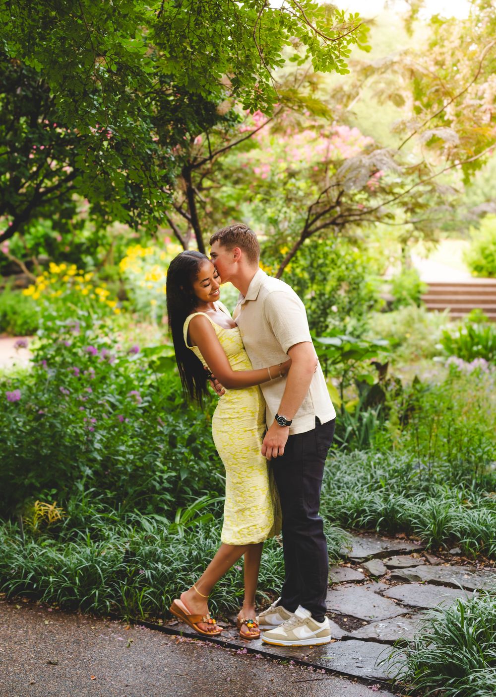Couple sharing romantic moments in a sunlit garden pathway surrounded by greenery.