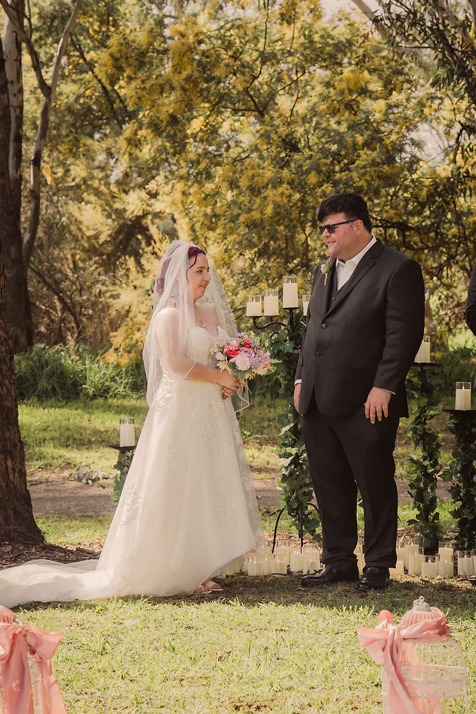 Bride and groom share a romantic moment in a sunlit outdoor setting with trees and pink accents.